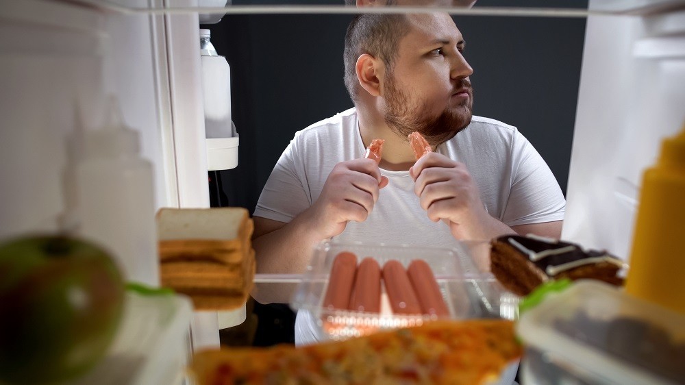 Hombre adicto a las comidas con hambre despues de haber cenado comiendo salchichas en secreto por la noche cerca del refrigerador, nutrición poco saludable